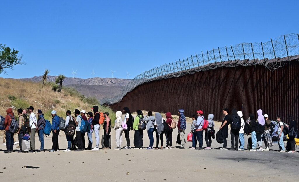 Migrantes esperan en fila con la esperanza de ser procesados ​​por los agentes de Aduanas y Patrulla Fronteriza después de que grupos llegaron a Jacumba Hot Springs, California, después de caminar bajo un intenso calor desde México a Estados Unidos el 5 de junio de 2024.